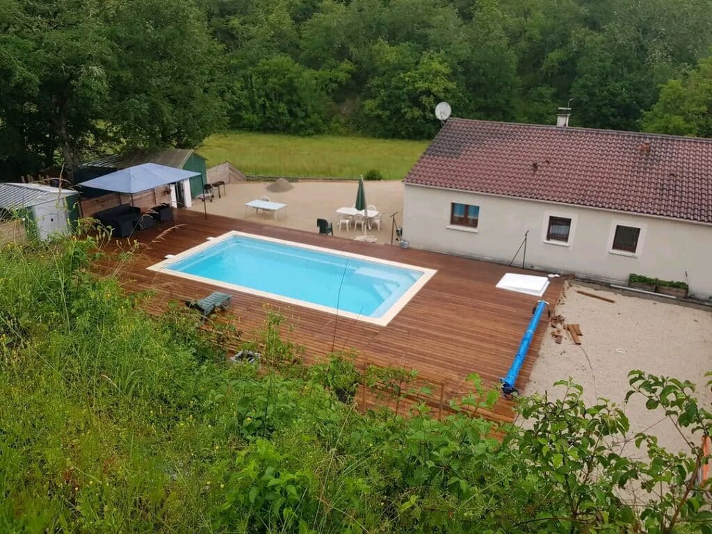Aménagement de jardin avec piscine et terrasse en bois Piscine rectangulaire avec grande terrasse en bois, mobilier de jardin et maison provençale entourée de verdure.