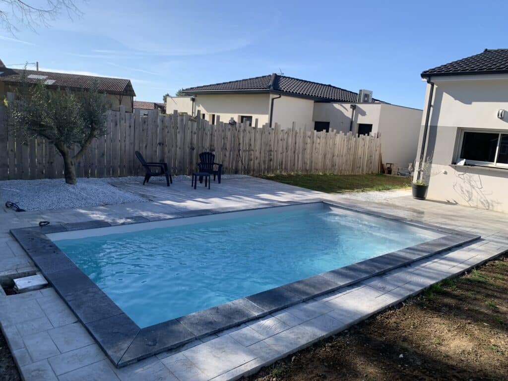 Aménagement piscine moderne avec terrasse et jardin Piscine rectangulaire moderne avec terrasse en dalles grises, clôture en bois et olivier dans un jardin ensoleillé.