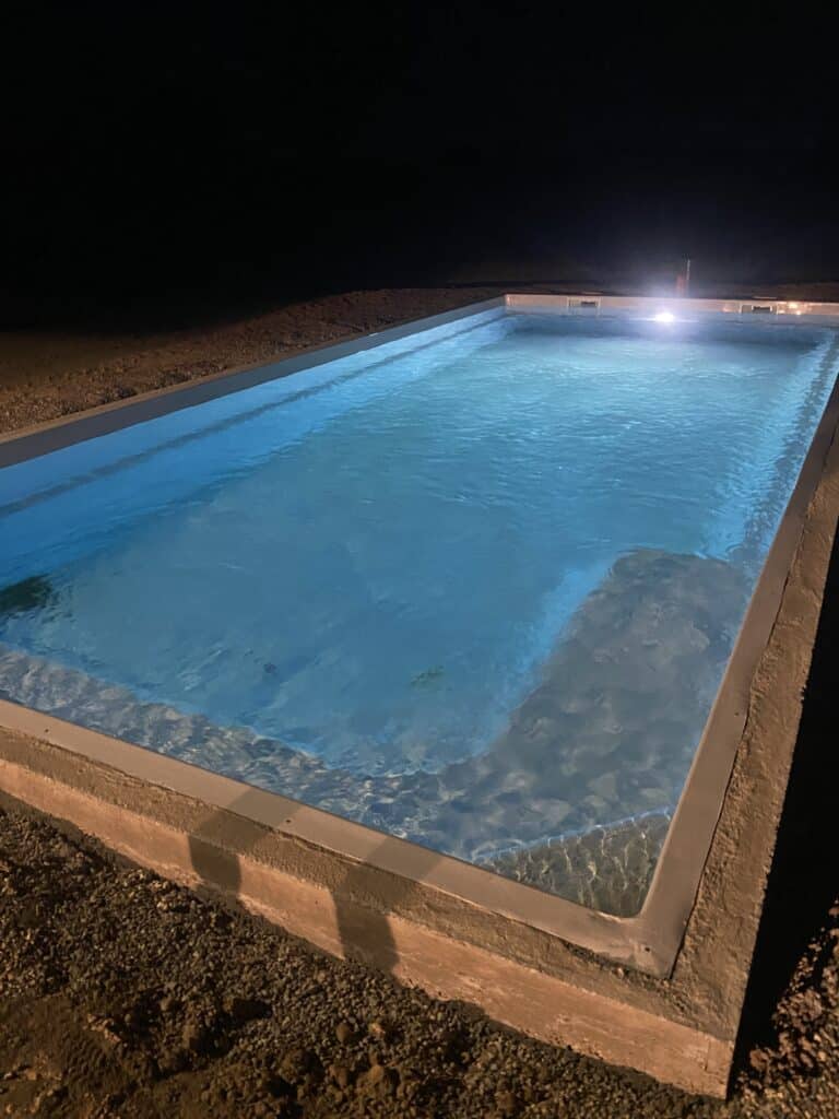 Piscine de Nuit avec Éclairage LED Bleu Subaquatique Piscine rectangulaire éclairée en bleu la nuit avec projecteur subaquatique et plage de gravier sombre.