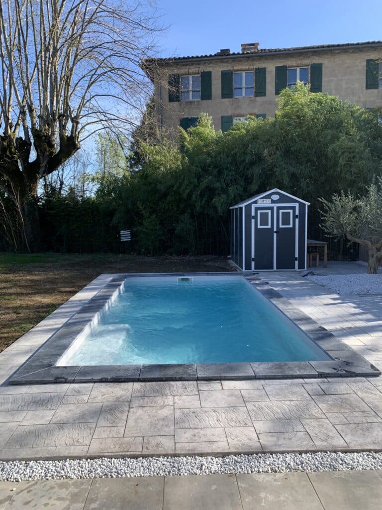 Piscine rectangulaire avec terrasse en pierre et jardin Piscine rectangulaire bleue, terrasse en dalles grises, abri de jardin PSW-533 et maison ancienne en arrière-plan.