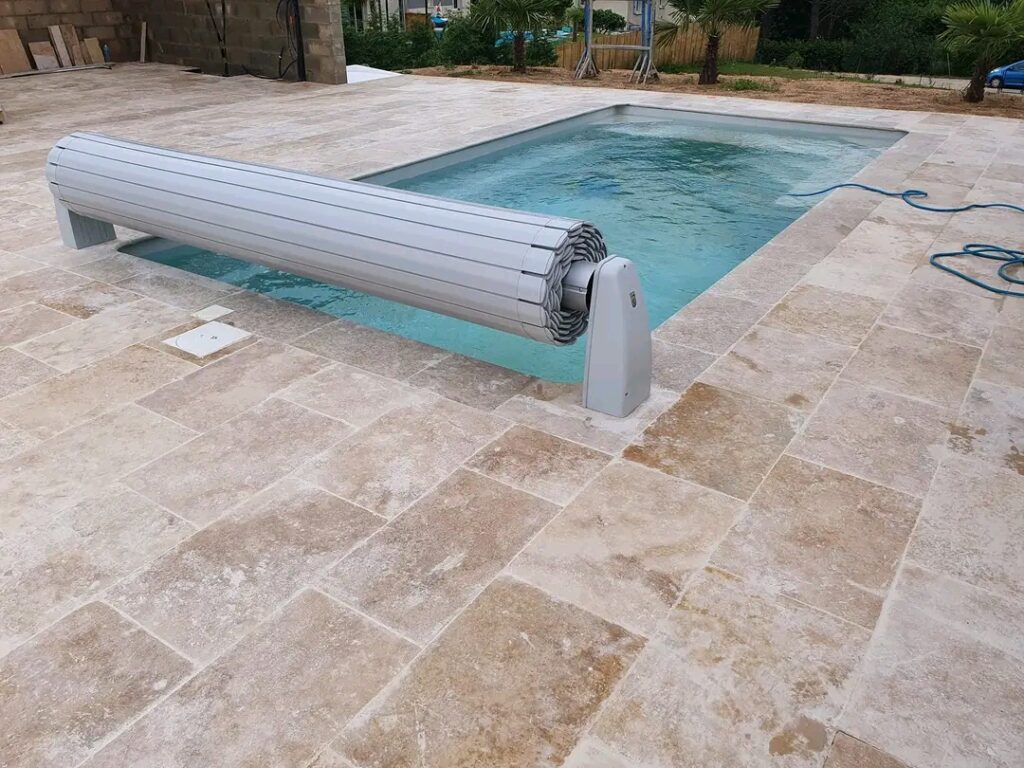 Volet de Piscine Automatique et Terrasse en Travertin Volet roulant automatique gris sur une piscine rectangulaire entourée de dalles de travertin beige clair en extérieur.